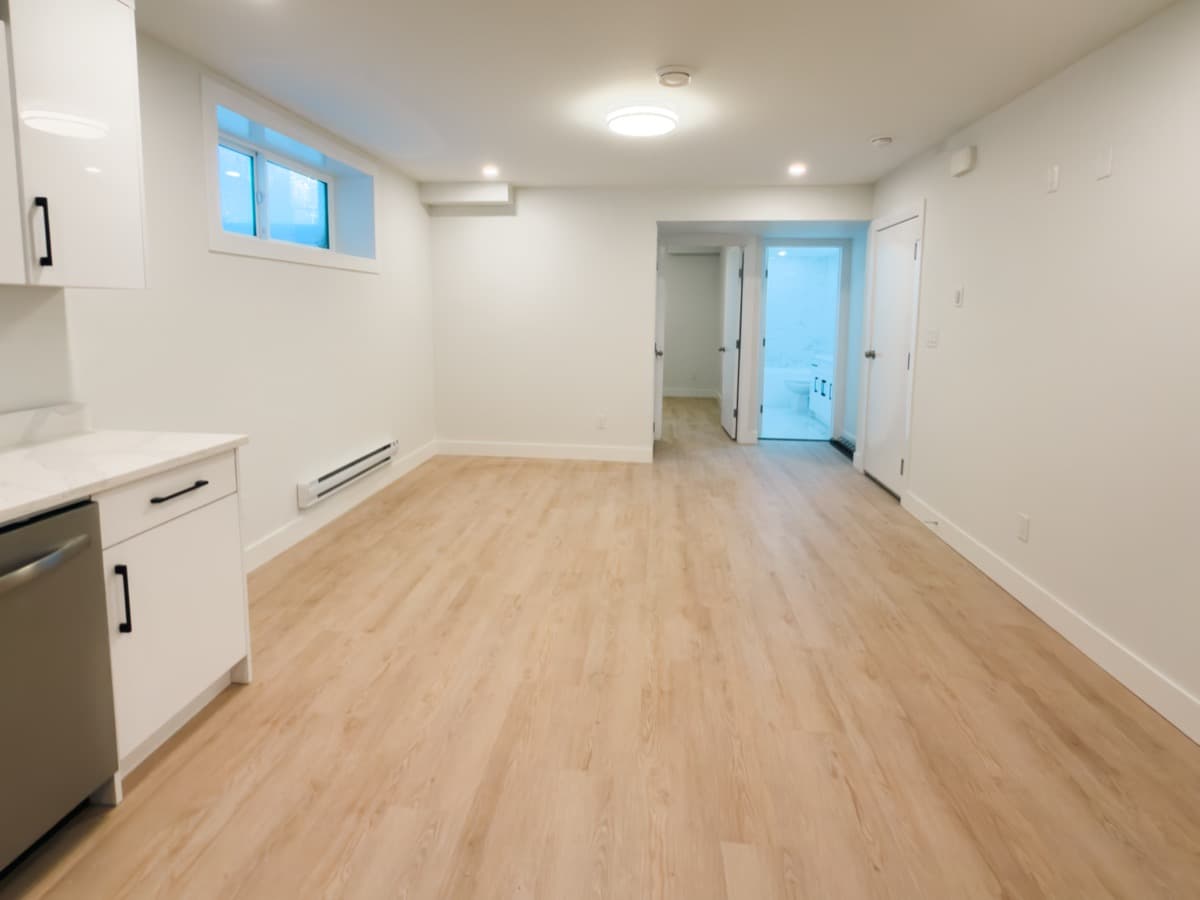 Finished basement living area with light plank flooring, a window, a baseboard heater and a hallway to the bathroom