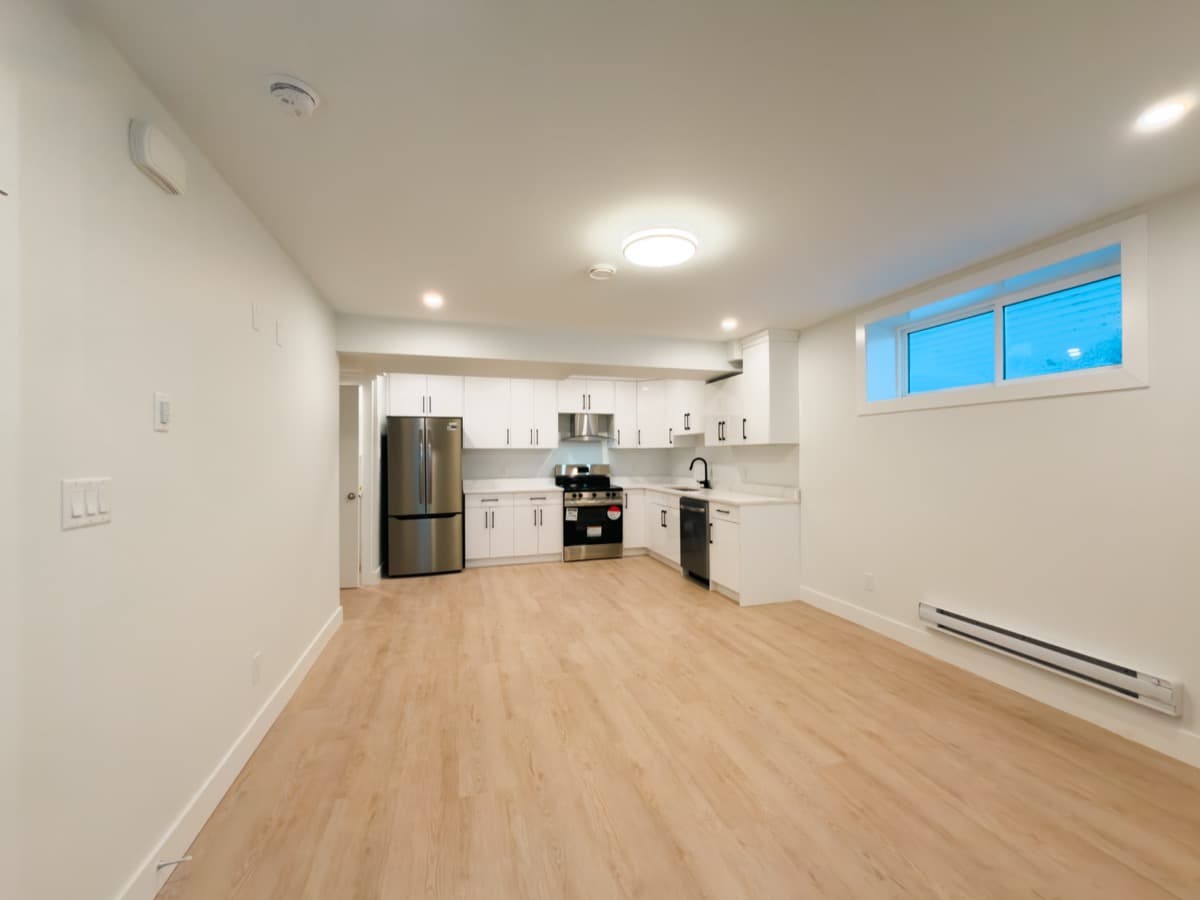 Long open room with light plank flooring leading to a white kitchen at the far end, below a basement window