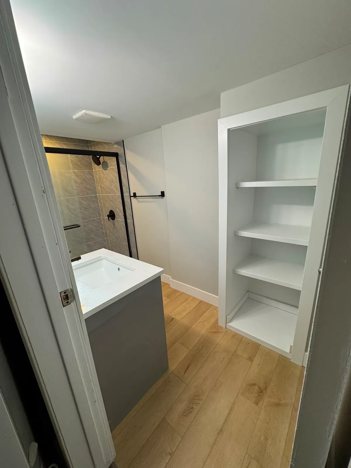 Built-in white shelving beside a grey vanity, with a black-framed shower door and wood-look plank floor behind