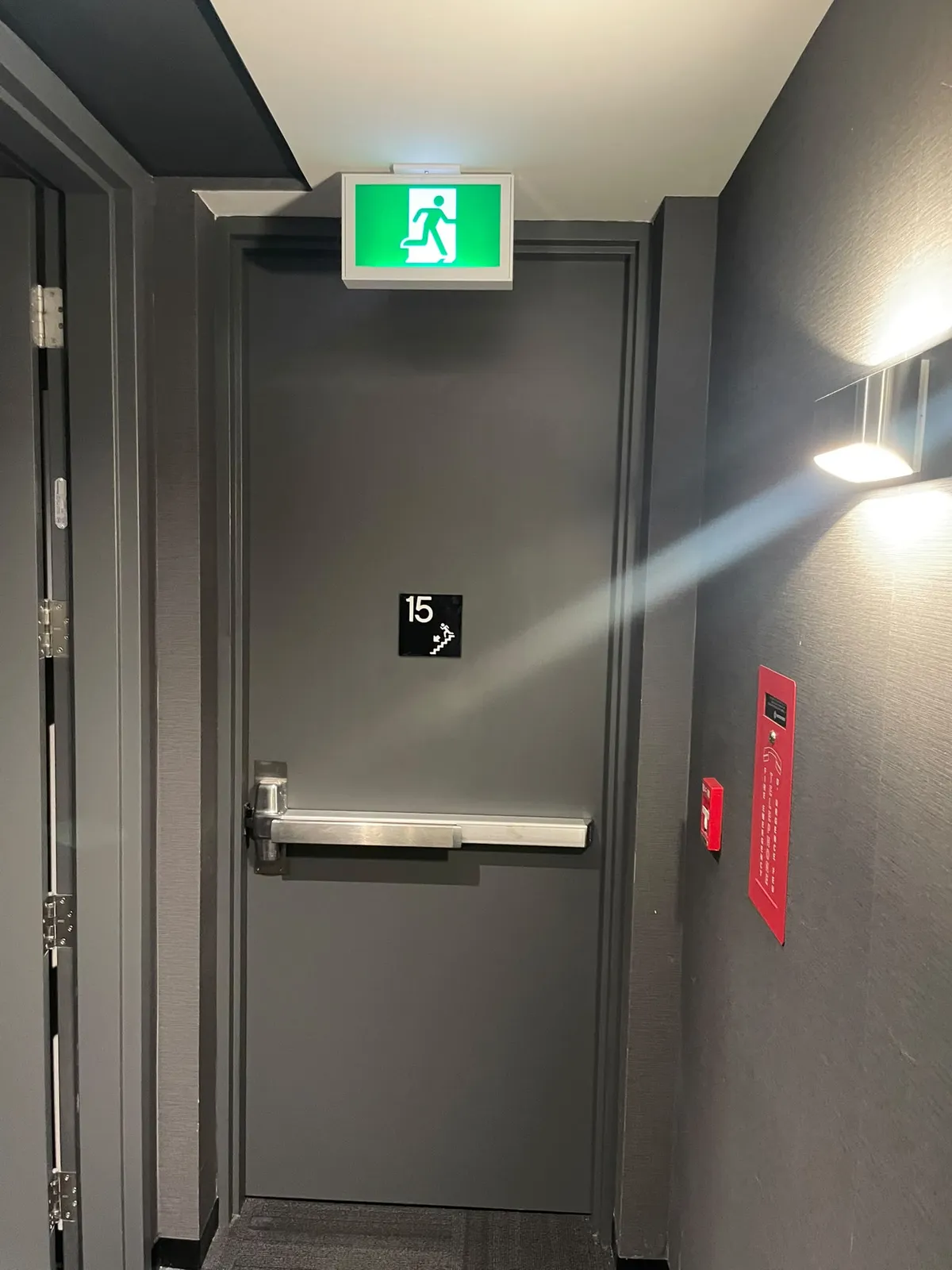 Grey exit door under a lit green running-man sign, with a wall sconce and a floor number beside the door