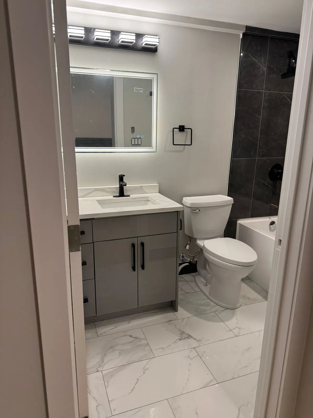 Suite bathroom with a grey vanity, a lit mirror, a toilet, dark wall tile by the tub and marble-look floor tile
