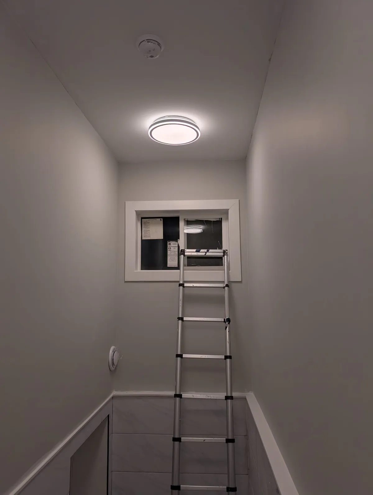 Narrow painted room with a window high on the end wall, a ceiling light and an aluminum ladder leaning up to the sill