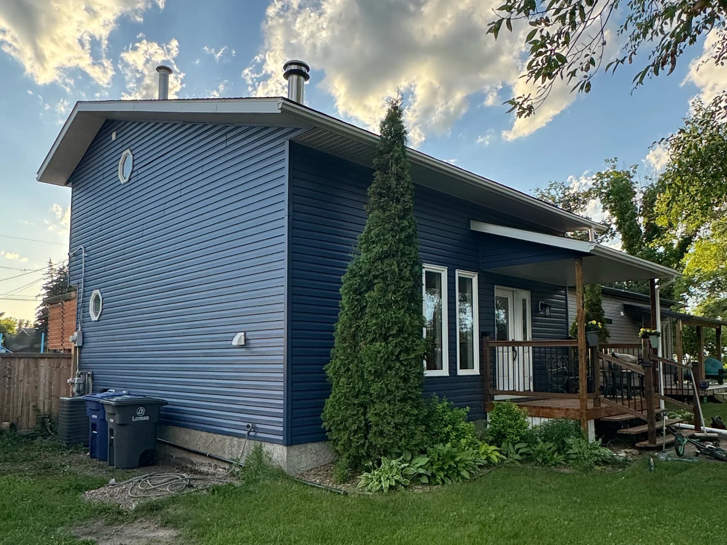 Side view of a house with new two-tone blue vinyl siding, a round gable window and a tall cedar by the porch